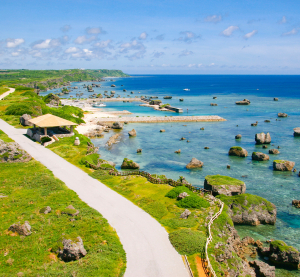 K&uuml;stenstra&szlig;e auf Okinawa mit Blick auf das t&uuml;rkisblaue Meer, ges&auml;umt von gr&uuml;nen Wiesen und verstreuten Felsen im Wasser.