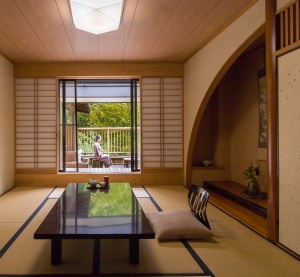 Traditionelles japanisches Ryokan-Zimmer mit Tatami-Matten, niedrigem Tisch und Blick auf eine Frau in Kimono auf der Terrasse.
