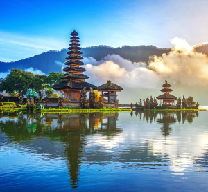 Traditioneller balinesischer Tempel am Seeufer inmitten tropischer Natur, mit Spiegelung im Wasser, umgeben von Nebelschwaden und grünen Bergen bei Sonnenaufgang.