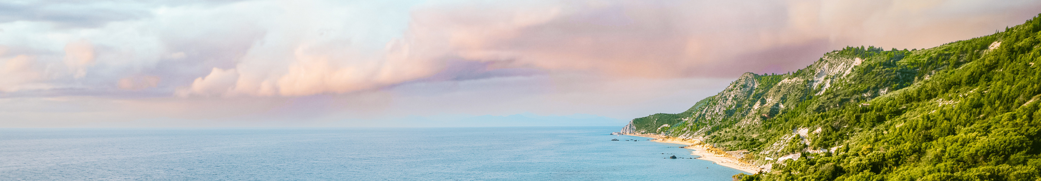 Blick auf das t&uuml;rkisfarbene Meer in Griechenland mit einem Sandstrand, gro&szlig;en Felsen, gr&uuml;nen H&uuml;geln im Hintergrund und Reihen von blauen Sonnenliegen am Ufer.