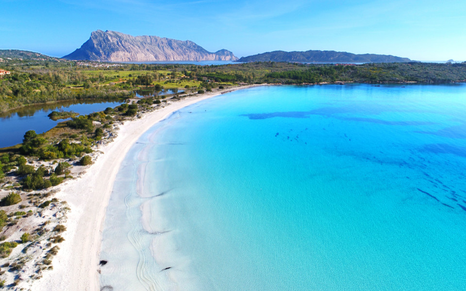 Blick auf einen Strand mit kristallblauem Meer und wei&szlig;em Sandstrand, dahinter ist ein Wald und Gebirge. 